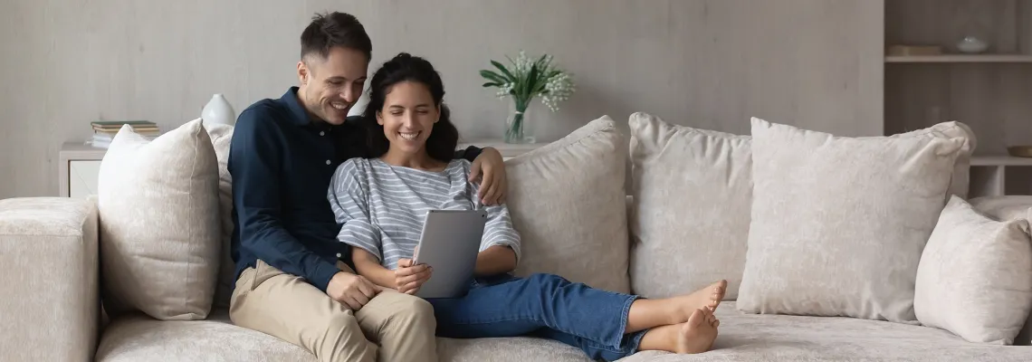 two people sit on a couch, happily looking together at a tablet